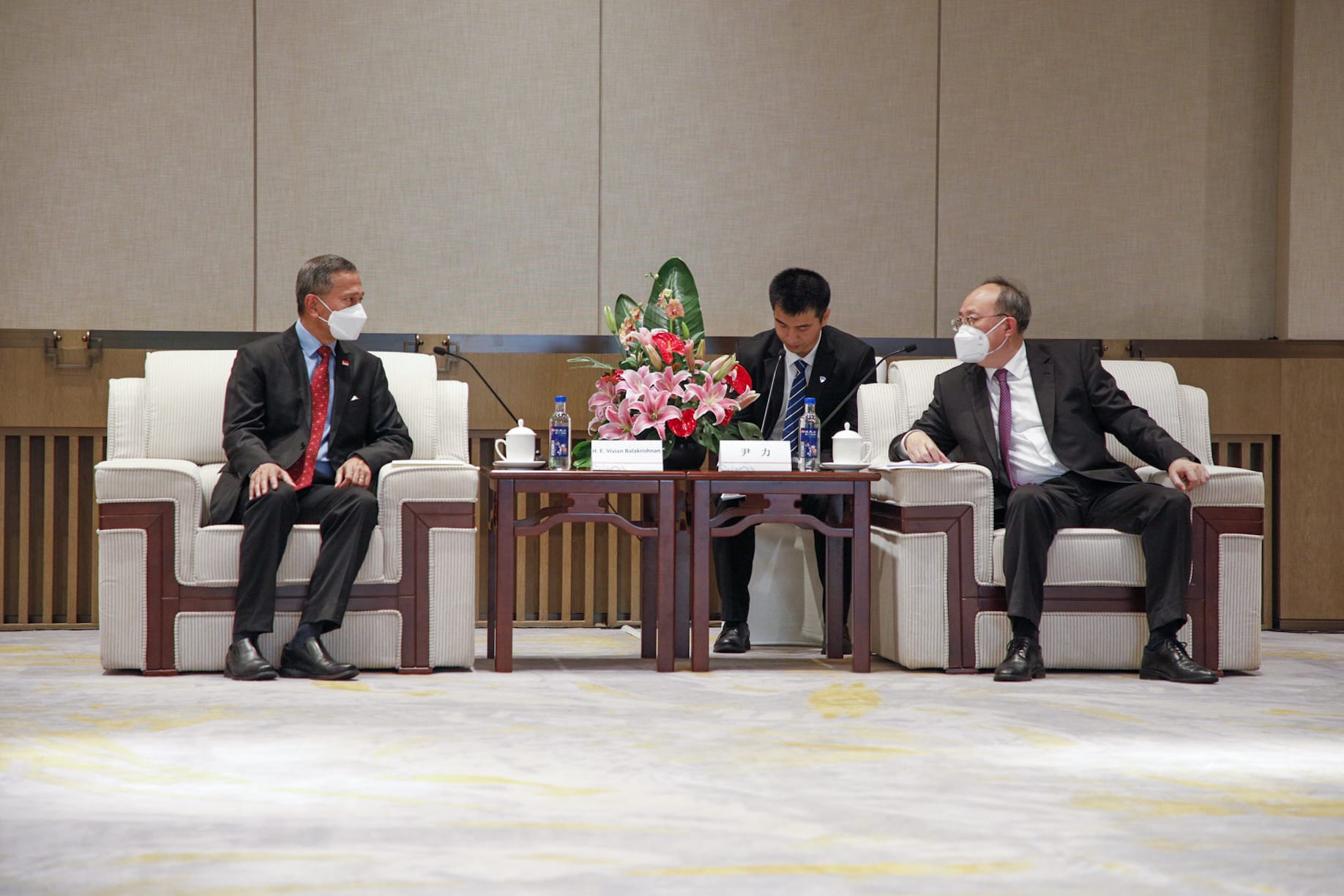 Two masked men in suits seated, flanking a man at a table with flowers, water.